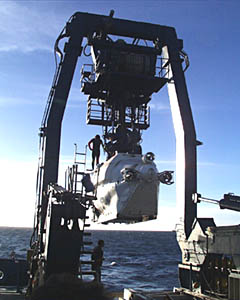 The A-frame swings out over the stern of the ship. The person standing on Alvin is one of two swimmers who will remove the lift lines when Alvin is in the water, freeing Alvin from the ship. 