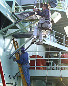  Jeff Benitz sits on a bosun&rsquo;s chair suspended from the ship&rsquo;s crane and chips paint from a rail. Jerry Graham paints a bulkhead below him.  
