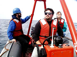  Jeff Benitz sits on a bosun&rsquo;s chair suspended from the ship&rsquo;s crane and chips paint from a rail. Jerry Graham paints a bulkhead below him.  