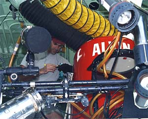 Bruce Strickrott doing maintenance on equipment in Alvin’s sail. The large black and yellow hoses going into the sail are air-conditioning and exhaust ducts to keep the electronic equipment cool and dry while on deck. 