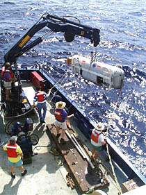 As the DSL-120 sonar vehicle is deployed off the port side of the fantail, its movements are controlled by quick release ropes attached at both ends of the instrument. These tag lines prevent the vehicle from hitting the side of the ship as it is lowered into the water. The vehicle is released from the small crane by a pull-pin release rope that Bob Elder is holding.