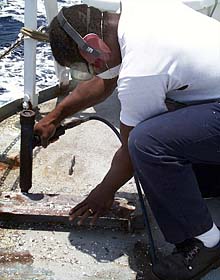 Cletus Finnel, an Ordinary Seaman (OS) on board R/V Melville is chipping rust off the deck using a needle gun. A needle gun is a pneumatic (air powered) tool that consists of a bunch of thick steel needles in a case. The needles vibrate very strongly so they chip away the surface layer of rust. Most people on the ship hate the needle guns because they make such a racket. People who have night watches have to learn to sleep through the loud noises which can't be avoided in the effort to keep the ship in good shape and free of rust. 