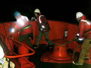 With one green line tied onto the net frame, Marine Science Technician Eddie Roggenstein loops it around the capstain (thick post) to hold it, while Jamee talks on the radio with the crane operator and Erich Horgan begins to attach another line.