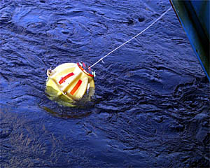 A transponder just after it is lowered into the water. The ship slowly moves away from it as the tether line is paid out. After all 185 meters of tether is paid out, the anchor is kicked over the side and the transponder sinks to the seafloor to start its work.