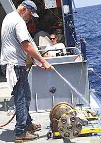 Bosun Bill Kamholtz washes down the main deck near the rock corer. We will use the rock corer later in the cruise to take samples of volcanic rocks produced by the new eruptions. Anja Marnewecke, the Third Mate, is taking a break after finishing her morning watch.