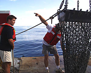 Ron Comer, the Scripps Resident Technician, gives hand signals to the A-frame operator during a lowering of the rock dredge today. Ben Wigham (left) is tending one of the safety lines used to keep the dredge from swinging as it is lifted over the side.