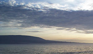 The classic “overturned soup bowl” profile of Galapagos volcanoes is well displayed by this early morning photo of Fernandina Island’s SW coastline. This particular type of volcanic profile is probably due to the fact that the volcanoes, once they emerge above sea level do not appear to have linear subaerial rift zones, like the volcanoes in Hawaii. Instead, they have radial rift zones, which form a bull’s eye pattern around the volcano. Eruptions along the radial rifts build up the slope concentrically around the center of the island and help build up the steep slope that marks the “bowl” part of the profile.