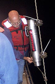 Gene Pillard, the Res. Tech., attaches the ‘pinger’ to the dredge wire. The pinger is attached 200 meters above the dredge bag. It sends a 12 kHz sound pulse to the seafloor every second. That pulse is received on the ship, as is the pulse that gets bounced off the seafloor. In the lab, a recorder measures the difference between the two acoustic returns from the pinger to give us the altitude of the pinger off the seafloor. This helps us determine if the dredge is really on the bottom collecting rocks.
