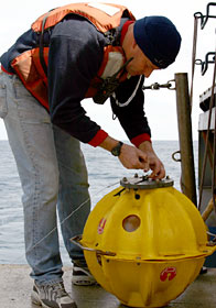 Mark Spear, a mechanical technician with the Alvin crew, prepares a transponder for dropping overboard. Transponders act as beacons to guide the submersible while exploring the seafloor. The hard yellow plastic case helps protect a glass ball, hydrophone, and computer panel inside.